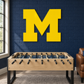 Michigan Block M yellow metal wall art above a foosball table in a navy brick game room.