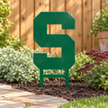 Michigan State Spartans block S metal yard art in green planted in a patio garden with buried stakes and a cedar fence.