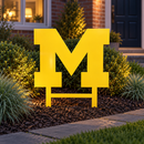 Michigan Wolverines metal yard art in yellow displayed in a landscaped front garden at dusk with buried stakes.