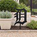 Old English D Metal Yard Art in Black with a buried center stake, displayed in a backyard baseball-practice garden.