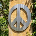 Full outdoor view of the peace symbol metal wall art hanging on a textured wall with natural sunlight and surrounding greenery.