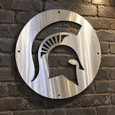 Circular Spartan helmet metal wall art featuring a clear coat finish mounted on a textured dark gray brick wall.