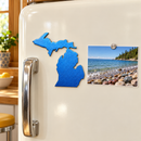 State of Michigan Metal Magnet in Metallic Blue, shown at its approximately 6-inch size on a refrigerator beside a lakeshore photograph.