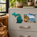 State of Michigan Metal Magnet in Metallic Green, shown at its approximately 6-inch size on a steel filing cabinet beside a lakeshore photograph.