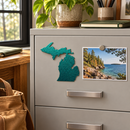 State of Michigan Metal Magnet in Metallic Green, shown at its approximately 6-inch size on a steel filing cabinet beside a lakeshore photograph.