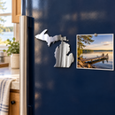 State of Michigan Metal Magnet in Polished Metal, shown at its approximately 6-inch size on a refrigerator beside a lakeshore photograph.